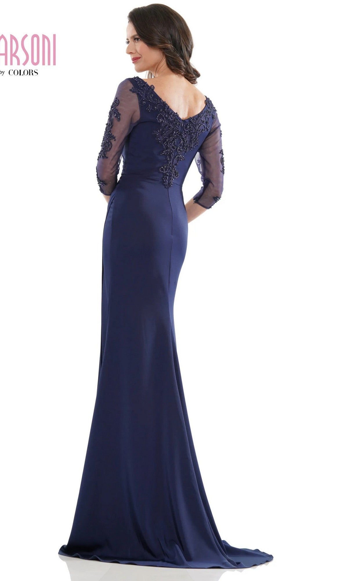 A woman stands facing away, wearing the Marsoni MV1146 long formal dress in navy blue, featuring a floor-length silhouette, embroidered sleeves, and lace detailing on the upper back.